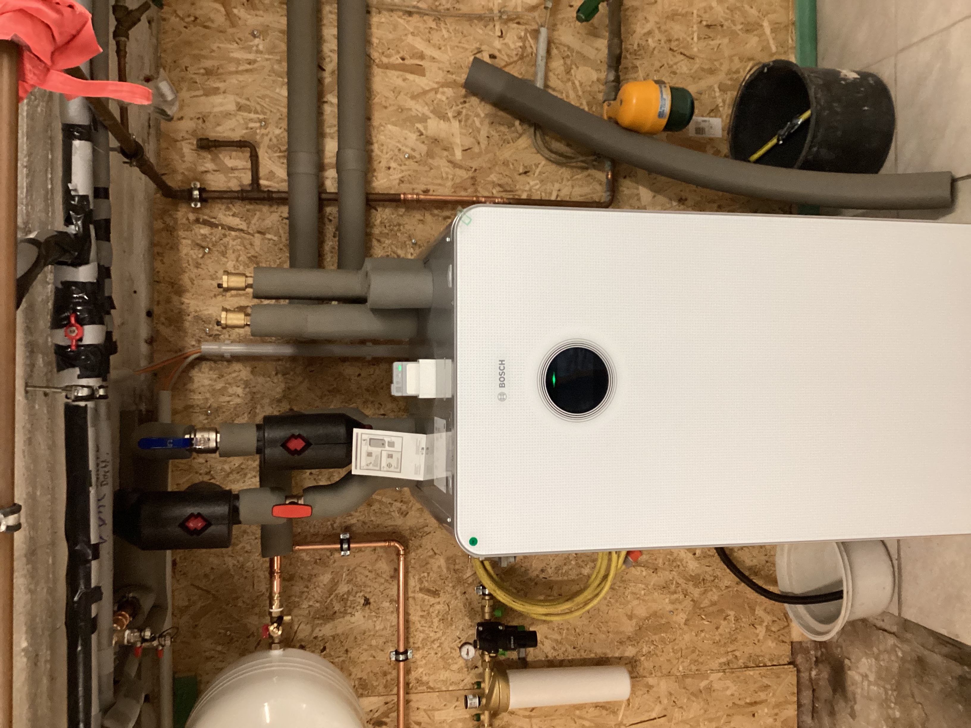 Eine weiße Bosch-Inneneinheit einer Wärmepumpe ist an einer OSB-Holzwand installiert, umgeben von gedämmten Leitungen und Technik.